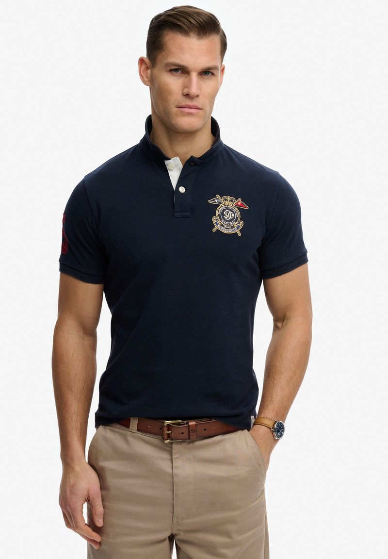 Mand iført marineblå polotrøje med broderet våbenskjold, beige bukser, brunt bælte og armbåndsur, stående med den ene hånd i lommen mod en hvid baggrund.