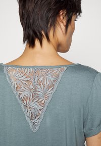 Haut court vert d'eau à manches courtes avec un dos en dentelle triangulaire présentant des motifs de feuilles et des bords festonnés, fabriqué à partir d'un matériau doux et léger.