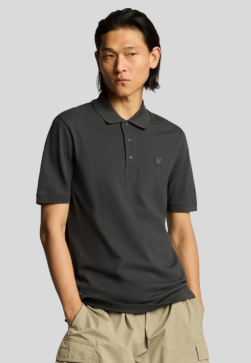 Polo shirt grigio scuro con colletto classico, maniche corte e un logo ricamato discreto sul lato sinistro del petto. Tessuto a trama liscia.