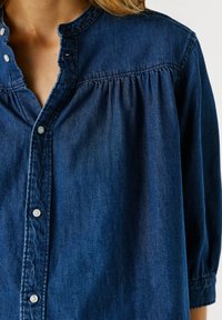 Chemise en denim bleu avec des boutons blancs, couture froncée à l'empiècement, et manches longues, portée partiellement déboutonnée au col.