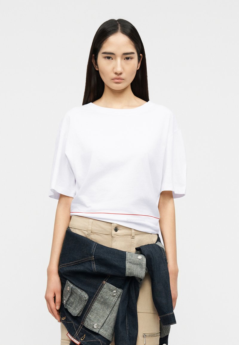 Νεαρή γυναίκα με μακριά σκούρα μαλλιά φοράει λευκό μπλουζάκι με κοντά μανίκια και κρατάει ένα σκούρο τζιν μπουφάν πάνω από μπεζ παντελόνι.