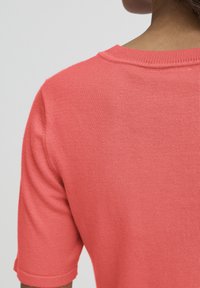 Pull corail à manches courtes avec un tricot texturé, un col rond et des poignets retroussés. Tissu lisse avec un léger éclat.