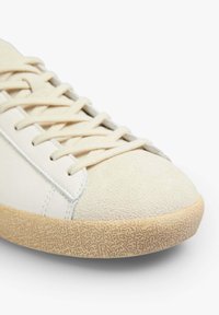 White leather sneaker with a textured suede toe, flat cotton laces, and a patterned gum rubber sole. Smooth finish with subtle stitching details.