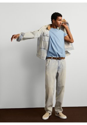 Un homme pose avec une veste en jean claire, un T-shirt bleu, un jean clair et des baskets couleur crème sur un fond de studio uni.