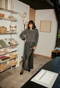 Cárdigan de punto gris con botones en el frente, combinado con una elegante falda maxi de satén negra. La modelo está de pie en un interior moderno con texturas naturales.