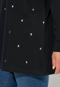 Pullover nero realizzato in morbido tessuto, con decorazioni a forma di teschio in argento distribuite uniformemente sulla parte anteriore. Fessure laterali all'orlo.