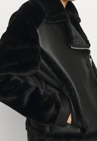 Svart fuskpäls- och läderjacka med bred krage och dragkedja, bärs av en person med handen i fickan, visad från axel till midja.