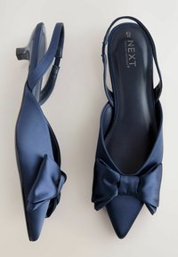 Tummansininen satiininen slingback-korkokenkä, jossa on terävä varvas ja kauniisti sotkeutuva rusetti, matalalla lohkareella ja sileällä tekstuurilla.
