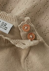 Beige neulevaate, jossa on aaltoileva kaulus, kaksi oranssia nappia ja teksturoitu kuvio. Hoito-ohjeet näkyvät reunassa.