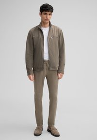 Khaki bomberjack met een ritsborstas, gecombineerd met bijpassende slim-fit broek. De stof lijkt glad; schoenen in een zachte, ingetogen tint.