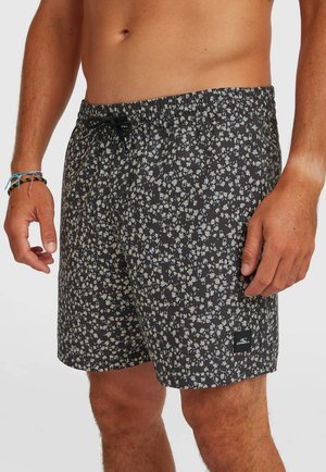 Mand iført sorte badeshorts med blomsterprint, elastisk talje og sort snøre, stående med hænderne afslappet ned langs siderne.