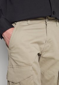 Man in zwart shirt met de linkerhand in de voorzak van beige cargobroek, met zichtbare knoop- en stikdetails.