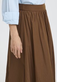 Main gauche d'une personne portant une chemise bleu clair aux manches retroussées, reposant à côté d'une jupe plissée marron avec une ceinture élastique.