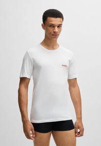 T-shirt en coton blanc avec le logo rouge "HUGO" sur la poitrine gauche, col rond, manches courtes et coupe standard. Porté avec un short noir.