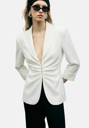 Donna che indossa un blazer bianco arricciato con maniche a tre quarti, pantaloni neri, fascia nera per capelli, grandi orecchini e occhiali da sole ovali neri.