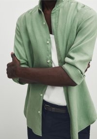 Grön linneskjorta med knappar och mandarin-krage, uppkavlade ärmar och knappar, bärs över en vit linne-topp, kombinerad med mörka byxor.