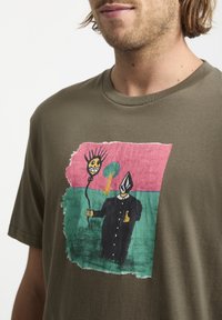 T-shirt en coton vert olive avec un imprimé graphique coloré représentant un personnage portant un chapeau pointu, un soleil et un arbre sur un fond texturé.