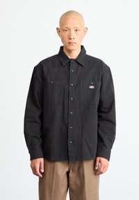 Black button-up shirt made of cotton with two chest pockets, contrasting buttons, and a small logo patch on the left pocket.