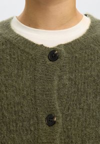 Olivgrön stickad kofta med en mjuk, fluffig textur, med rund halsringning och tre svarta knappar fram.