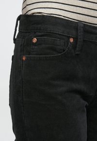 Juodos spalvos korduro kelnės su vertikaliais grioveliais, puoštos vario spalvos kniedėmis, diržo kilpomis ir šonine kišene, matomos nuo juosmens žemyn.