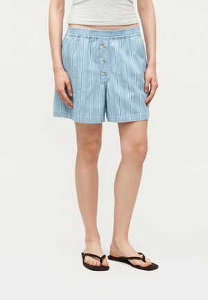 Άτομο που φοράει ανοιχτό μπλε ριγέ σορτς με κουμπιά μπροστά, λευκό κομμένο τοπ και μαύρα σανδάλια τύπου σαγιονάρα, στέκεται μπροστά σε απλό φόντο.