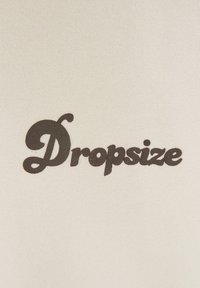Weißer Stoff mit einem dunkelbraunen "Dropsize"-Text in einer stilisierten Kursive, der die glatte Textur mit einem matten Finish betont.