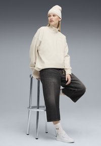 Helle beigefarbene Jacke mit Reißverschluss, dunkle cropped Jeans, weiße Sneakers mit Logo, weiße Socken, beiger Beanie, sitzt auf einem Metallhocker.