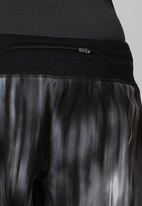 Gros plan sur un short de sport noir et gris avec une ceinture noire et une petite poche zippée horizontale.