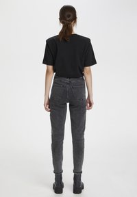 Camiseta negra combinada con jeans negros desgastados de tiro alto que cuentan con bolsillos traseros. La modelo lleva botas negras hasta el tobillo, vista desde atrás.