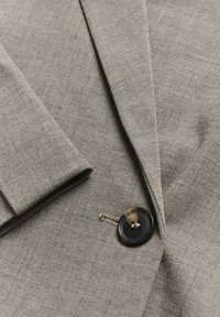Svetlošedý blazer s textúrovanou tkaninou, s výstrihom do tvaru V, ostrými klopami a okrúhlym kontrastným hnedým gombíkom.