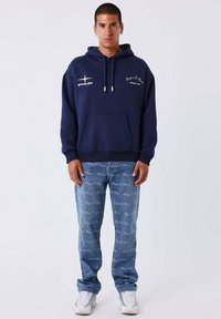 Marineblauwe hoodie van katoen met een kangoeroezak, voorzien van witte logo-tekst en zilverkleurige accenten. Gepaard met lichtblauwe, gemusterde jeans.