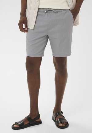 Mand iført lysegrå shorts med snor, sorte sandaler, cremefarvet skjorte og stribet åben knap-skjorte, stående med hænderne i lommerne.
