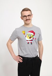 Grå kortærmet t-shirt med et farverigt grafisk motiv af en julesvamp SpongeBob SquarePants, der holder en klokke. Sort bælte, sorte bukser.
