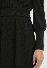 Czarna sukienka z teksturowanej tkaniny o subtelnym połysku, dopasowana w talii, z bufiastymi rękawami, tworząca zwiewną sylwetkę.