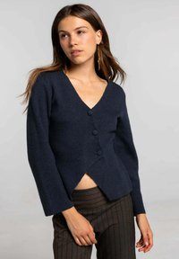 Cárdigan de punto azul marino con cuello en V, tres botones y bajo asimétrico. Presenta mangas largas y una textura suave. Se ven pantalones a rayas.