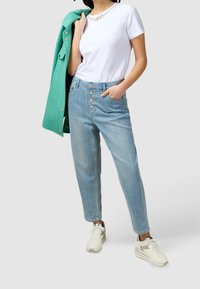 Žena v bílém tričku, světle modrých vysokých džínech s dekoračními knoflíky, v bílých teniskách, držící zelenou bundu přes rameno.