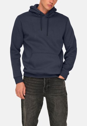 Uomo che indossa una felpa con cappuccio blu navy con tasca frontale e coulisse, abbinata a jeans grigio scuro su uno sfondo semplice.