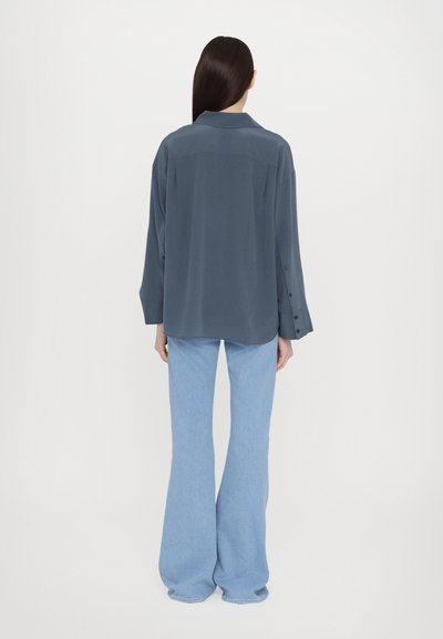 Blusa grigia a maniche lunghe con colletto, polsini abbottonati e vestibilità morbida, abbinata a jeans a zampa d'elefante blu chiaro. Texture e forme semplici.