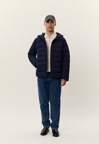 Νεαρός άνδρας με ναυτικό μπουφάν puffer, μπεζ πουλόβερ με φερμουάρ στον γιακά, μπλε τζιν, μαύρα παπούτσια και γκρι καπέλο μπέιζμπολ, στέκεται μπροστά σε απλό φόντο.