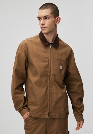 Brązowa kurtka zapinana na zamek z kołnierzem typu corduroy, długimi rękawami i kieszenią na piersi z małą naszywką z logo. Miękka, teksturowana tkanina.