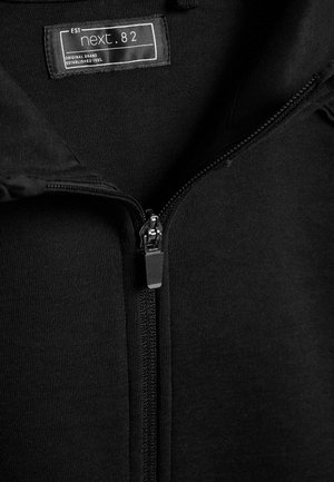 Must musta zipiga kapuutsiga, mis on valmistatud pehmest kangast, keskel on metallist tõmblukk ja silt, millel on bränd "next. 82."