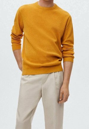 Mand iført sennepsgul langærmet striktrøje og beige plisserede bukser, stående med den ene hånd i lommen foran en ensfarvet baggrund.