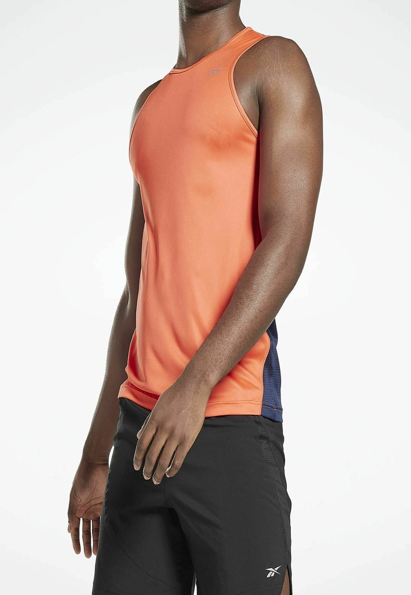 Homme portant un haut de sport sans manches orange avec des panneaux latéraux bleu marine et un short noir avec un petit logo blanc, debout devant un fond uni.