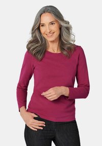 Fuchsia Langarmoberteil mit Rundhalsausschnitt, aus weichem Stoff, kombiniert mit dunklen Jeans. Das Model posiert mit den Händen in den Hüften und lächelt.