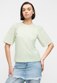 Νέα γυναίκα με ίσια σκουρόχρωμα μαλλιά φοράει ανοιχτό πράσινο τοπ με φουσκωτά μανίκια και σκισμένη denim φούστα πάνω από τζιν, με φόντο λευκό.