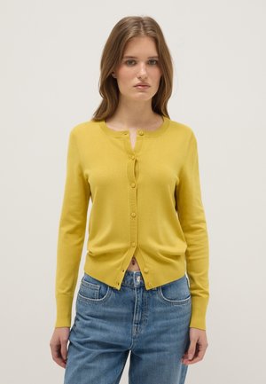 Vrouw die een gele, lange mouwen, met knopen voorzien cardigan en blauwe spijkerbroek draagt, staand tegen een effen lichte achtergrond.