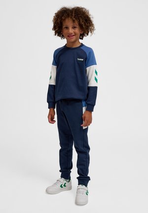 Marineblauwe sweatshirt met witte mouwen en groene accenten, gecombineerd met marineblauwe sweatpants. Heeft een logo op de borst en witte sneakers.