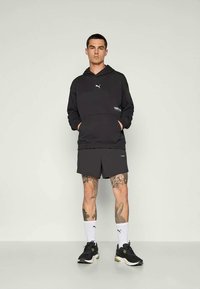 Felpa con cappuccio nera con logo, pantaloni corti scuri e sneaker neri. Calze bianche con logo esposte su uno sfondo neutro.
