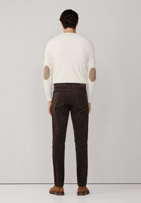 Hombre de pie con la espalda hacia la cámara, vistiendo un suéter blanco con parches marrón en los codos, pantalones marrón oscuro y zapatos marrón contra un fondo plano.