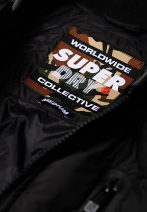 Veste noire avec un patch camouflage à l'intérieur portant la mention "Worldwide Superdry Collective" et une petite étiquette de taille "Medium" en dessous.